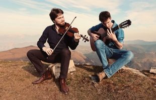 Anton Jablokov and Stefano Moccetti sitting on rocks on top of a mountain playing their instruments.